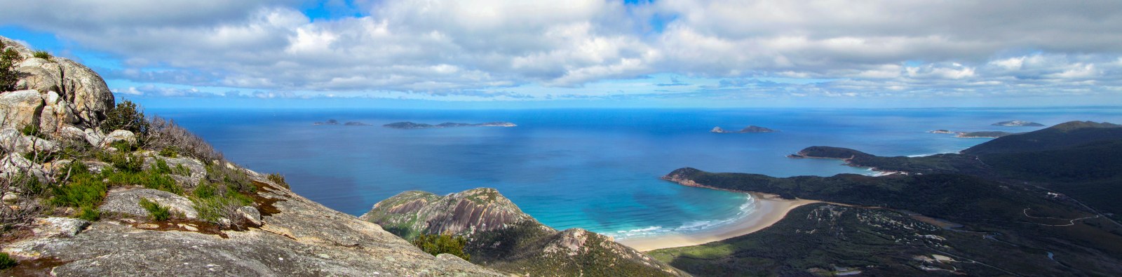 Turquoise bay with a curved white beach seen from a rocky summit, small islands on the horizon