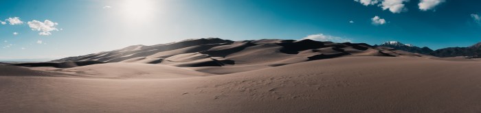 Rolling sand dunes stretching toward distant mountains under a bright sky with scattered clouds