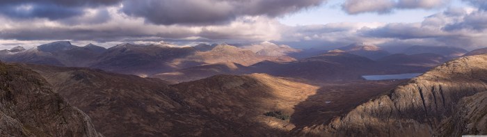 Panorama of brown Scottish Highland mountains and glens under heavy clouds, with a small loch in the valley