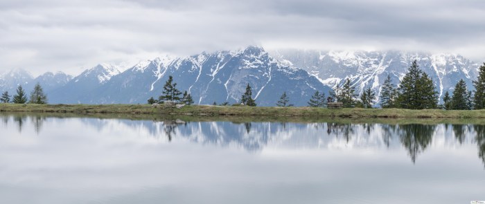 Snowy Austrian mountains and pine trees reflected in a still lake under grey clouds