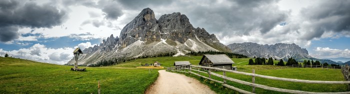 Gravel path through green alpine meadows toward a jagged Dolomites peak, with wooden huts and a wayside cross