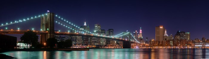 Brooklyn Bridge strung with lights at night, with the Manhattan skyline reflected in the East River